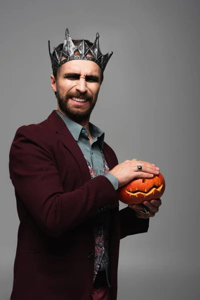 Scared man in vampire king halloween crown covering creepy pumpkin with hand isolated on grey — Stock Photo