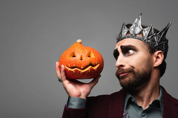 Hombre reflexivo en el rey vampiro corona de Halloween mirando calabaza tallada aislada en gris - foto de stock