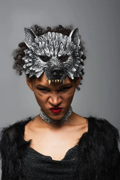 Furiosa mujer afroamericana en hombre lobo máscara de Halloween frunciendo el ceño a la cámara aislada en gris - foto de stock