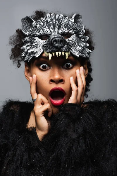 Femme afro-américaine choquée dans le masque de loup-garou halloween touchant visage isolé sur gris — Photo de stock