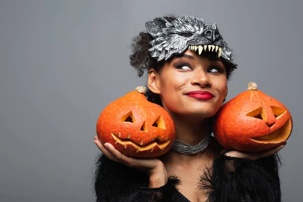 Mujer afroamericana complacida en hombre lobo máscara de Halloween sosteniendo calabazas talladas aisladas en gris - foto de stock