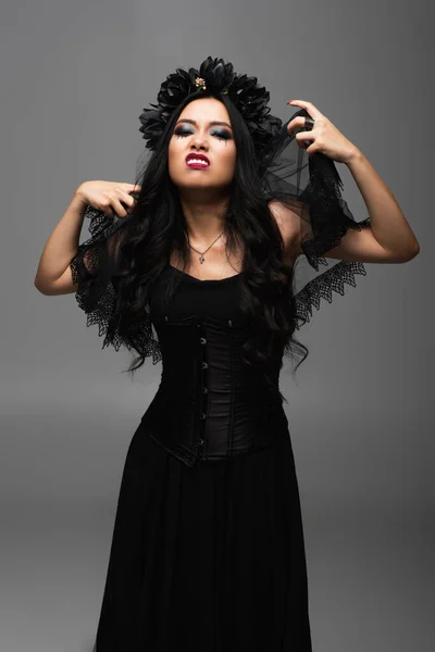 Asian woman in vampire halloween costume touching black veil and grimacing isolated on grey — Stock Photo