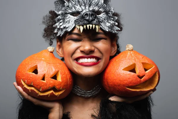 Overjoyed african american woman in wolf halloween mask holding carved pumpkins and smiling with closed eyes isolated on grey — Stock Photo