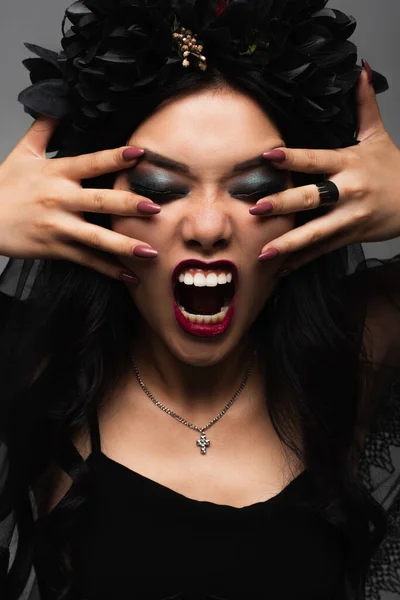 Asian woman in vampire halloween makeup touching face and growling with closed eyes isolated on grey — Stock Photo