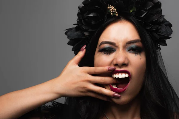 Страшная азиатка в костюме вампира Хэллоуина держит руку у открытого рта, изолированного на сером — стоковое фото