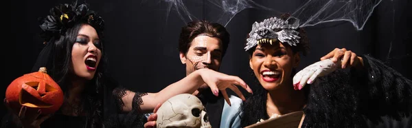 Fröhliche multiethnische Freunde in Halloween-Kostümen haben Spaß mit gruseligem Totenkopf und Spielzeughand auf schwarz, Banner — Stockfoto