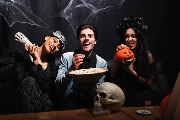 Happy multiethnic friends with popcorn, toy hand and halloween pumpkin looking at camera near spooky skull on black — Stock Photo