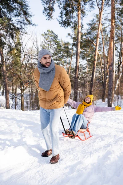 Homme souriant tirant traîneau avec petite amie dans le parc d'hiver — Photo de stock