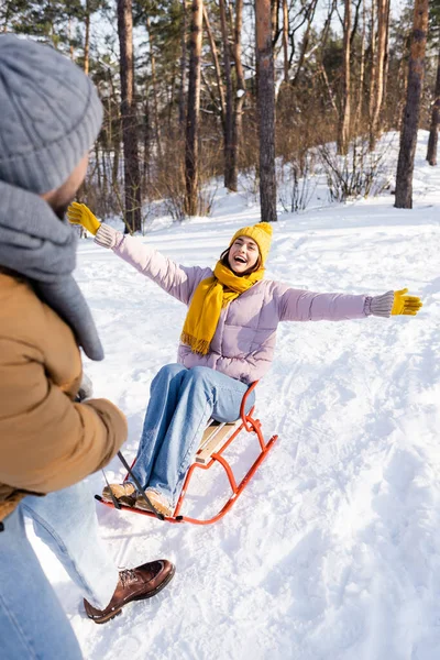 Mulher alegre sentado no trenó perto namorado borrado no parque de inverno — Fotografia de Stock