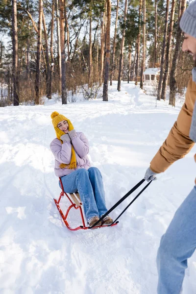 Mulher alegre em luvas e chapéu sentado no trenó perto namorado borrado no parque de inverno — Fotografia de Stock