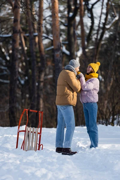 Casal alegre em luvas de mãos dadas perto de trenó no parque de inverno — Fotografia de Stock