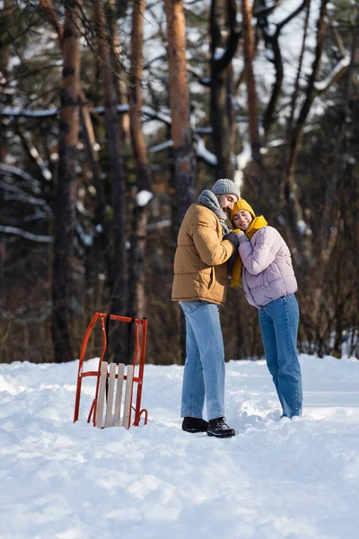 Jeune couple souriant tout en se tenant la main près du traîneau dans le parc d'hiver — Photo de stock