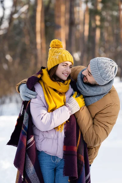 Uomo sorridente abbracciando fidanzata con coperta nel parco invernale — Foto stock