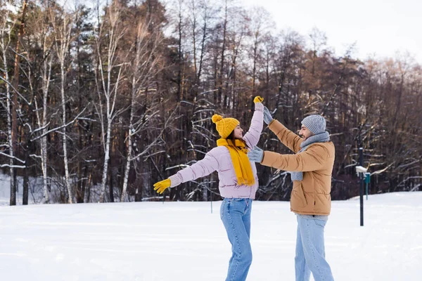 Vista lateral de pareja positiva en sombreros calientes tomados de la mano y divirtiéndose en el parque de invierno - foto de stock