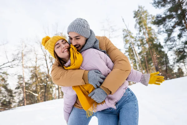 Захоплений чоловік у в'язаному капелюсі, що обіймає дівчину із закритими очима в парку зі снігом — стокове фото
