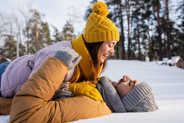 Vista lateral da mulher positiva em chapéu de malha deitado no namorado na neve no parque — Fotografia de Stock