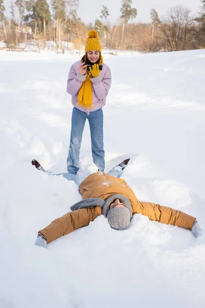 Lächelnde Frau macht Foto, während Freund Schnee-Engel im Park macht — Stockfoto