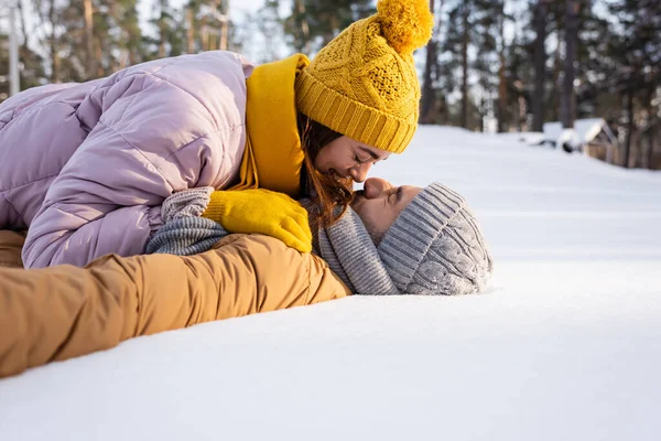 Вид збоку молодої пари в капелюхах цілується, лежачи на снігу — стокове фото