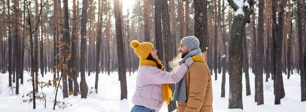 Side view of cheerful woman embracing boyfriend in hat and scarf in winter park, banner — Stock Photo