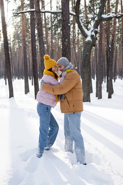Вид сбоку улыбающейся пары в зимнем наряде, обнимающейся в снежном парке — стоковое фото