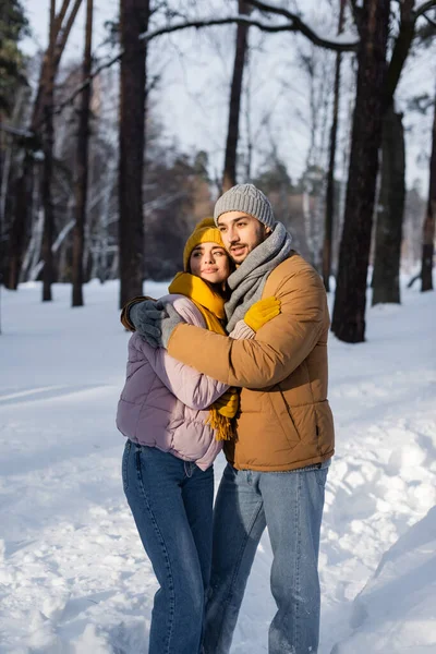 Jeune couple en tenue d'hiver embrassant tout en se tenant debout dans la lumière du soleil et la neige — Photo de stock