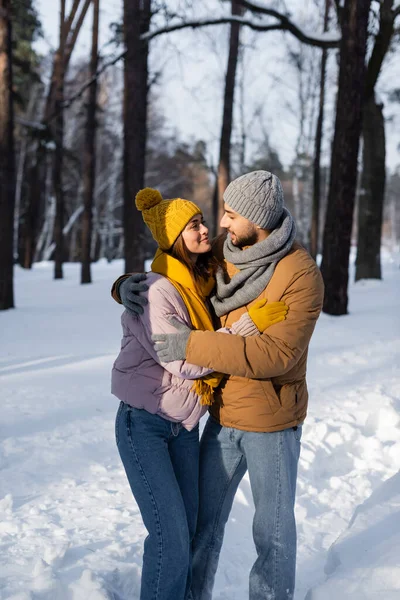 Улыбающаяся пара в теплой одежде, смотрящая друг на друга, обнимаясь в зимнем парке — стоковое фото