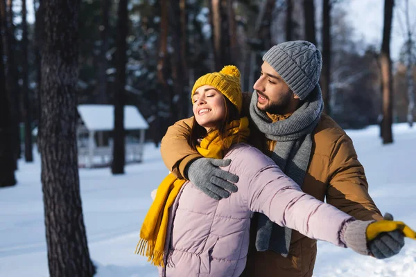 Hombre abrazando y cogido de la mano de la novia feliz con los ojos cerrados en el parque de invierno - foto de stock