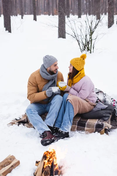 Seitenansicht eines lächelnden Paares mit Tassen am Lagerfeuer im Winterpark — Stockfoto