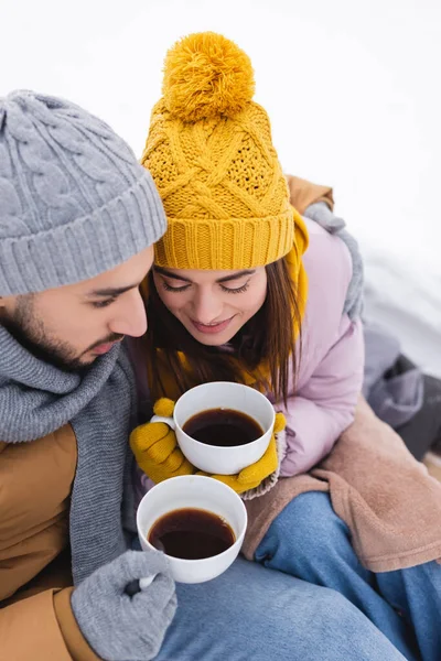 Высокий угол обзора пары в зимнем наряде держа кофейные чашки в снежном парке — стоковое фото