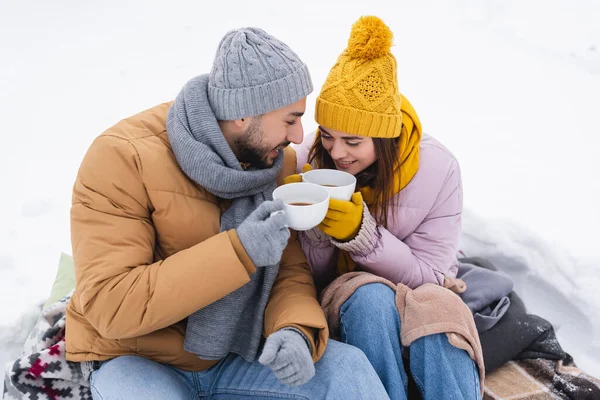 Pareja positiva sosteniendo tazas de café sentado en mantas en la nieve - foto de stock