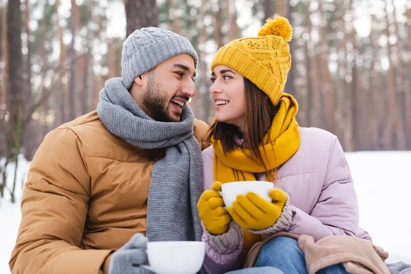 Hombre feliz en sombrero de punto sosteniendo taza cerca de la novia en el parque durante el invierno - foto de stock