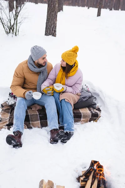 Couple avec tasses à café tenant la main près de feu de joie dans un parc enneigé — Photo de stock