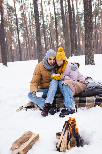 Uomo abbracciare fidanzata con tazza vicino falò offuscata sulla neve — Foto stock