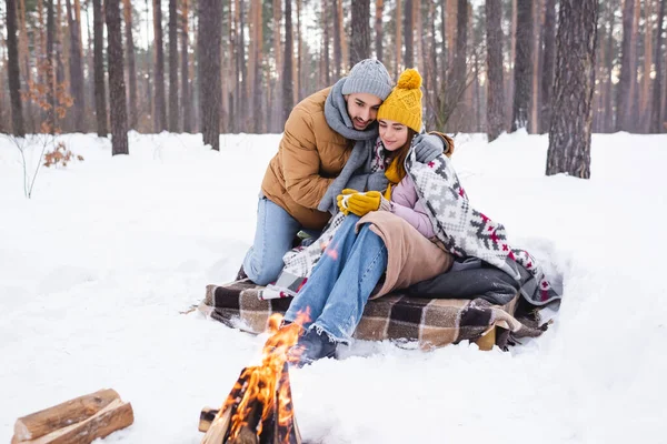 Uomo abbracciare fidanzata con tazza vicino falò nel parco invernale — Foto stock