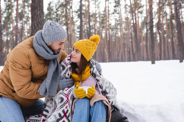 Homem feliz abraçando namorada bonita em chapéu de malha e cobertor segurando copo no parque de inverno — Fotografia de Stock