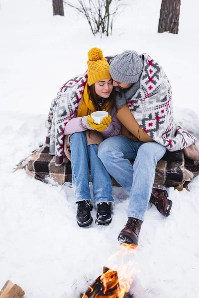 Junge Frau im Winteroutfit und Decke hält Tasse neben Freund und Lagerfeuer im Schnee — Stockfoto