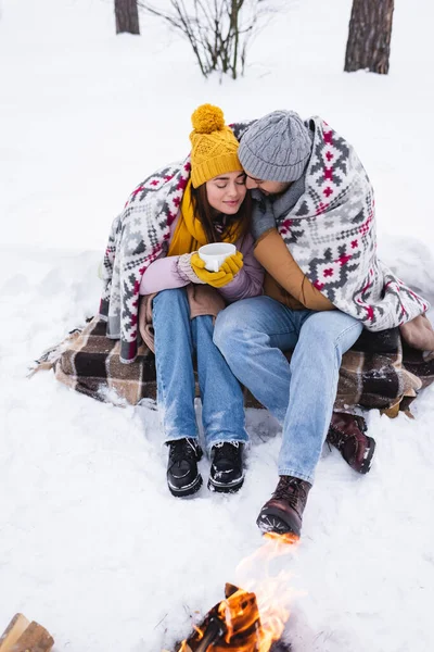 Giovane donna in abito invernale e coperta in possesso di tazza vicino fidanzato e falò sulla neve — Stock Photo