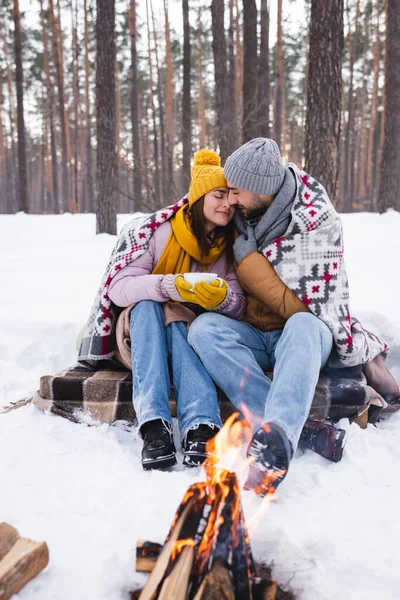 Jovem casal com cobertor e copo de aquecimento perto de fogueira borrada no parque de inverno — Fotografia de Stock
