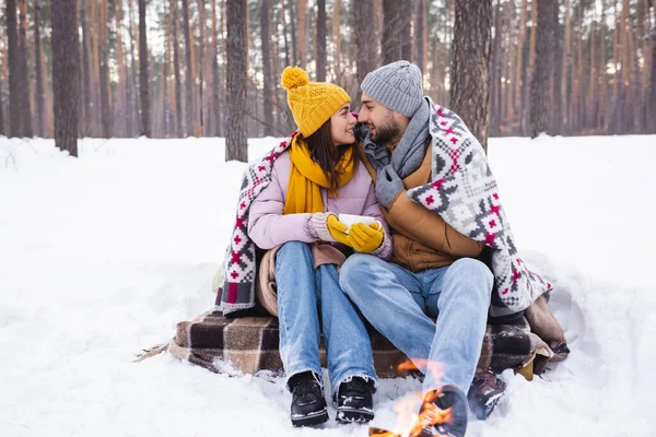 Side view of positive couple with cup and blanket looking at each other near blurred bonfire in winter park — Stock Photo