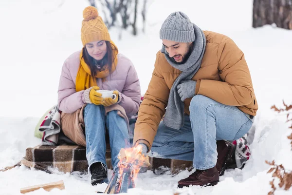 Sonriente hombre sosteniendo tronco cerca de hoguera y novia con taza en el parque de invierno - foto de stock