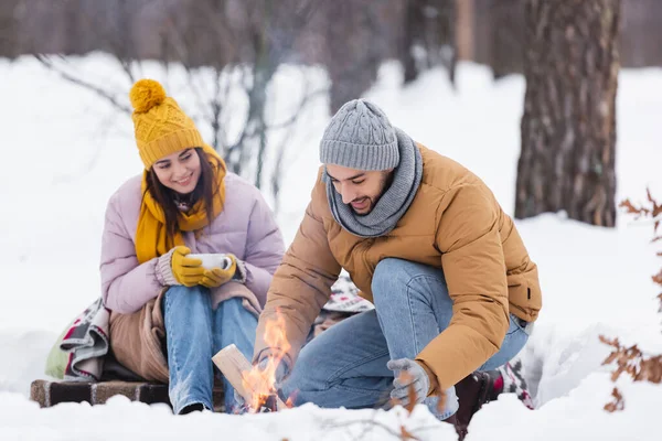 Fröhlicher Mann hält Baumstamm am Lagerfeuer und Freundin mit Tasse im verschneiten Park — Stockfoto
