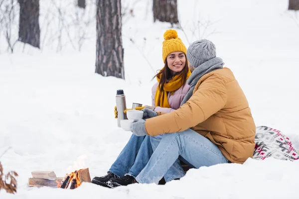 Homme tenant des tasses près de petite amie souriante avec thermos et feu de joie dans un parc enneigé — Photo de stock