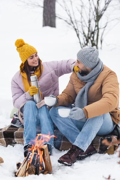 Fröhliche Frau hält Thermoskanne neben Freund mit Tassen und verschwommenem Lagerfeuer im Winterpark — Stockfoto