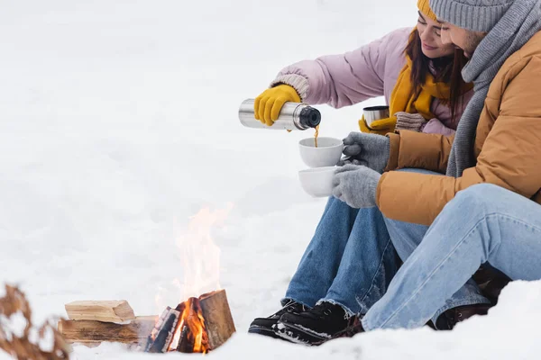 Woman pouring coffee from thermos near boyfriend and bonfire in winter park — Stock Photo