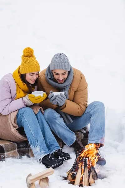 Couple with cups looking at bonfire on snow in park — Stock Photo