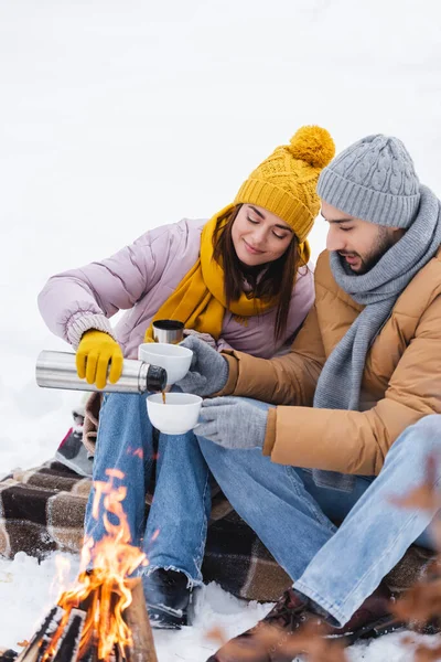 Homem segurando copos perto da namorada com garrafa térmica e fogueira borrada no parque de inverno — Fotografia de Stock