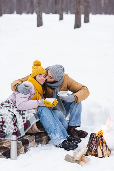 Homem beijando namorada com copo e cobertor perto de fogueira no parque de inverno — Fotografia de Stock