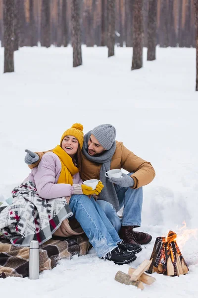 Smiling man pointing with finger near girlfriend with cup and bonfire in winter park — Stock Photo
