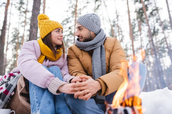 Pareja joven mirándose mientras se calientan las manos cerca de una hoguera borrosa en el parque de invierno - foto de stock