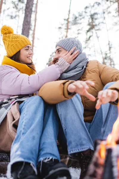 Visão de ângulo baixo de mulher sorridente ajustando chapéu de malha de namorado perto fogueira borrada no parque de inverno — Fotografia de Stock
